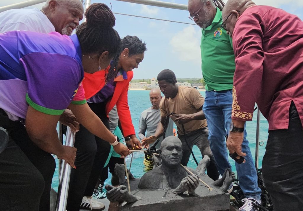 photo of a group of people grouped around a scuplture of a head and five sets of clasped hands on a boat with ropes attached to the scupture